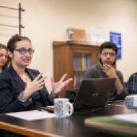 students seated around a table listening to their professor speak